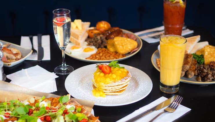 A table set with various breakfast dishes, including pancakes topped with fruit, an egg and sausage platter, toast, baked beans, and corn. Drinks like orange juice, a Bloody Mary, and a glass with a fruit garnish accompany the meal.