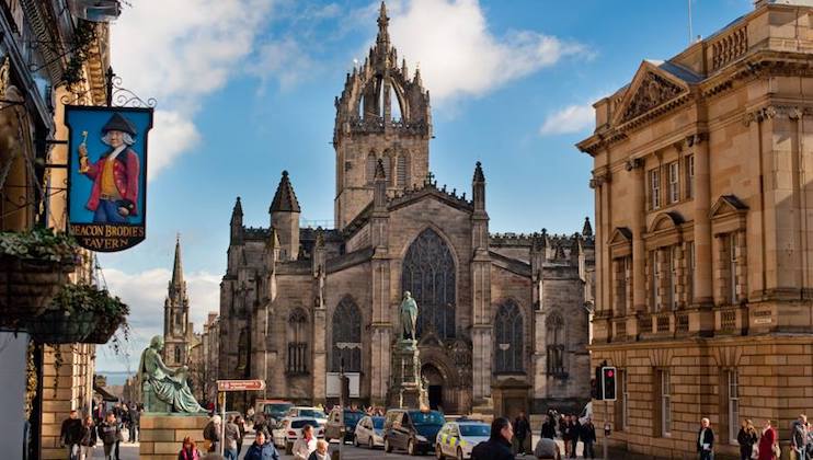 A bustling street scene featuring a historic Gothic church with a tall spire at the center. To the left, there is a pub sign for Deacon Brodie's Tavern. People walk along the street, flanked by old stone buildings under a bright blue sky with scattered clouds.