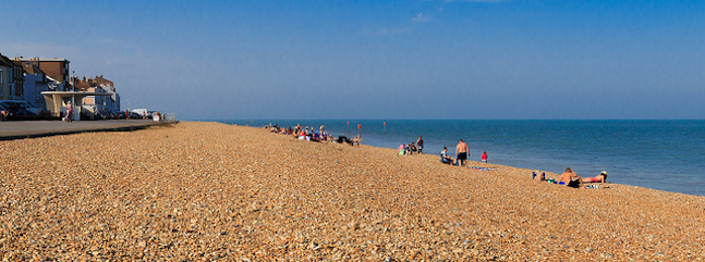 A scenic beach with a rocky shoreline stretches into the distance. People are sitting and sunbathing near the water. A row of seaside buildings can be seen on the left, and the clear blue sky meets the calm sea on the right.