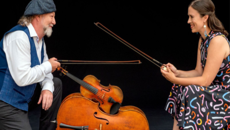 A man with a gray beard, wearing a blue vest, white shirt, and a gray beret, and a woman in a colorful dress sit facing each other. They hold violin bows and smile warmly, with a cello and a violin placed between them against a dark background.