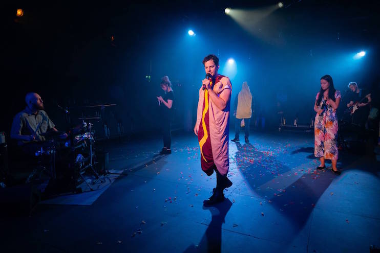 A performer wearing a hot dog costume and holding a microphone stands center stage under blue lighting. Other performers, including a drummer and musicians, are also on stage. Confetti is scattered on the ground.