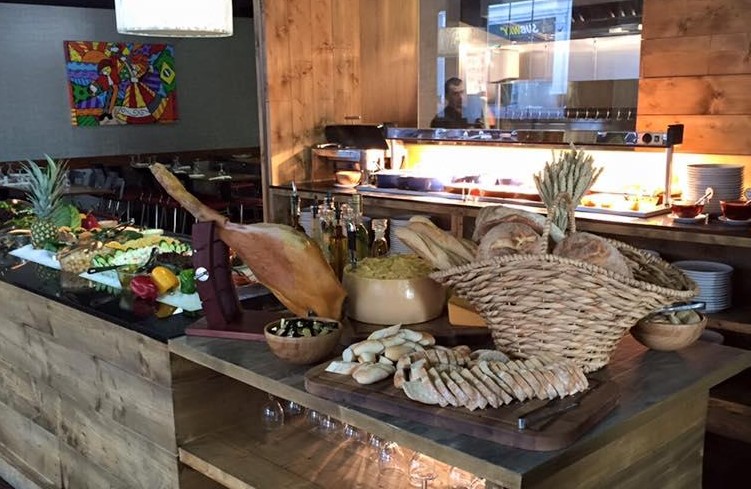 A buffet spread in a rustic restaurant features various breads, breadsticks, and fresh fruit, including pineapples. A basket overflows with loaves, and a cured ham sits on a stand. An assortment of oils, nuts, and spreads is also available. Artwork on the wall enhances the ambiance.