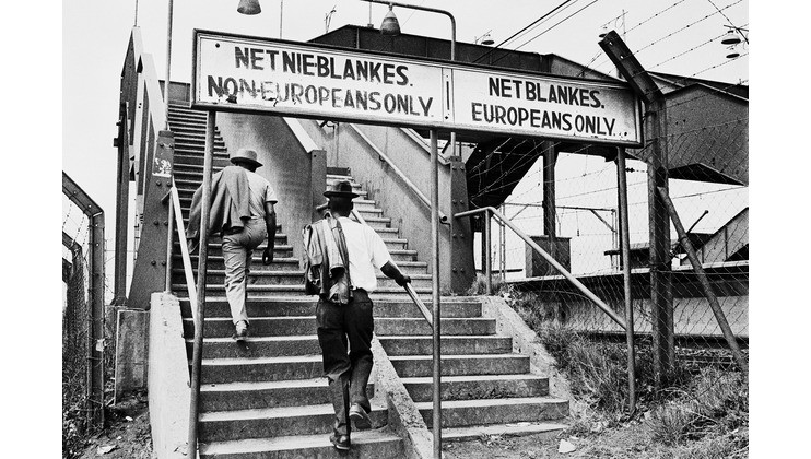 A black-and-white photograph showing two men walking up separate staircases divided by signs. The left sign reads NET NIE-BLANKES. NON-EUROPEANS ONLY and the right sign reads NET BLANKES. EUROPEANS ONLY, highlighting segregation during apartheid in South Africa.