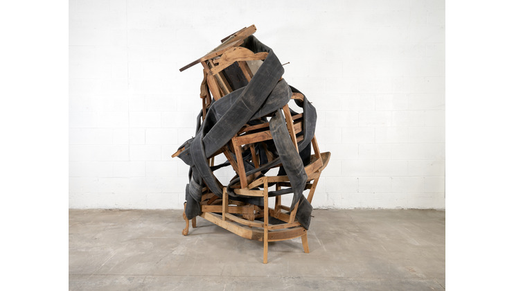 A sculpture composed of wooden chairs and benches intricately stacked and intertwined, wrapped in a dark, leathery material. The structure stands against a plain white brick wall on a concrete floor, creating a contrast between the organic forms and industrial setting.