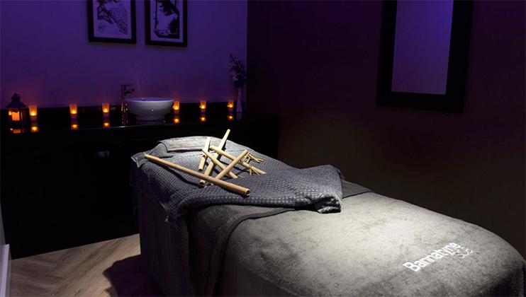 A dimly lit massage room featuring a massage table draped with a dark grey towel and bamboo sticks placed on top. Candles line a shelf in the background, creating a serene atmosphere. A sink and framed pictures are also visible on the back wall.