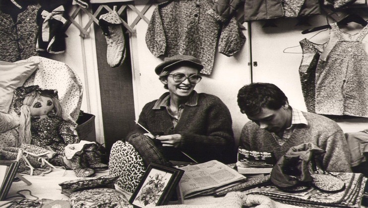 Black-and-white photo of two people at a market stall. One person is knitting and smiling, wearing glasses and a cap. The other is looking down at a notebook. Various fabrics, garments, and a doll are displayed around them. The ambiance is cheerful and busy.