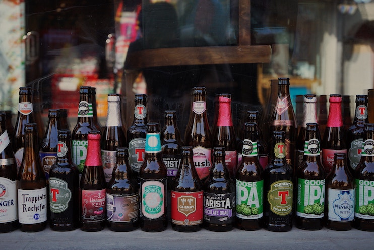 A display of various beer bottles is arranged in front of a shop window. The bottles showcase different brands and types of beer, including lagers, ales, and IPAs, with colorful labels and various bottle shapes and sizes prominently visible.