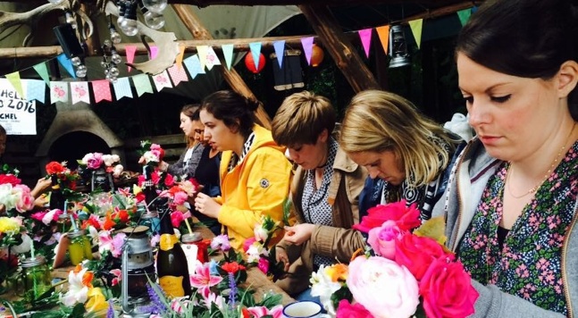 A group of people are seated at a long table, arranging flowers into bouquets. The table is filled with various colorful flowers and crafting supplies. Bunting and decorations hang above them in an outdoor setting. Everyone appears focused on their work.