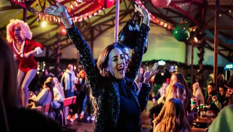 A joyful woman with raised arms dances at a lively indoor party filled with people. The venue is decorated with colorful overhead lights and balloons. In the background, a performer in vibrant costume entertains on a stage. Attendees are seated and standing.