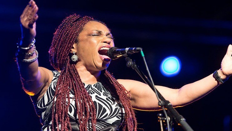A woman with long braided hair passionately sings into a microphone on stage, with her arms outstretched. She is wearing a patterned sleeveless top, and is illuminated by stage lighting, including a blue spotlight in the background.