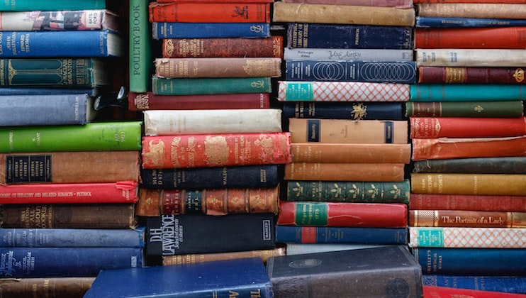 A cluttered bookshelf filled with a variety of colorful, old and new books stacked both vertically and horizontally. The books come in various sizes and colors, with some titles visible and others obscured by the arrangement.