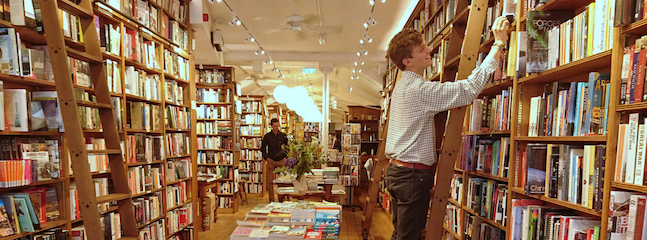 A crowded bookstore with tall wooden shelves filled with books. A person on the left scans the shelves, while a person on the right stands on a ladder, reaching for a book on an upper shelf. The center has a table displaying more books. The ceiling has spotlights.