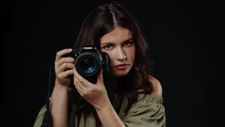 A woman with long brown hair holds a Canon DSLR camera up to her face, as if ready to take a photo. She is wearing an off-the-shoulder green top and is set against a dark background. Her expression is focused and intense.