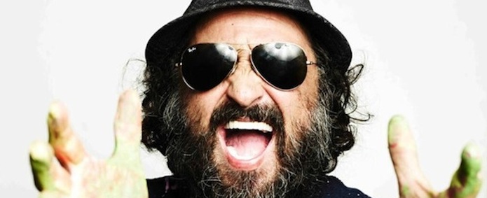 A man with long curly hair and a thick beard wearing a black hat and aviator sunglasses is enthusiastically facing the camera with his mouth wide open. His hands are raised and seem to be covered in paint, with a vivid expression of excitement or joy.