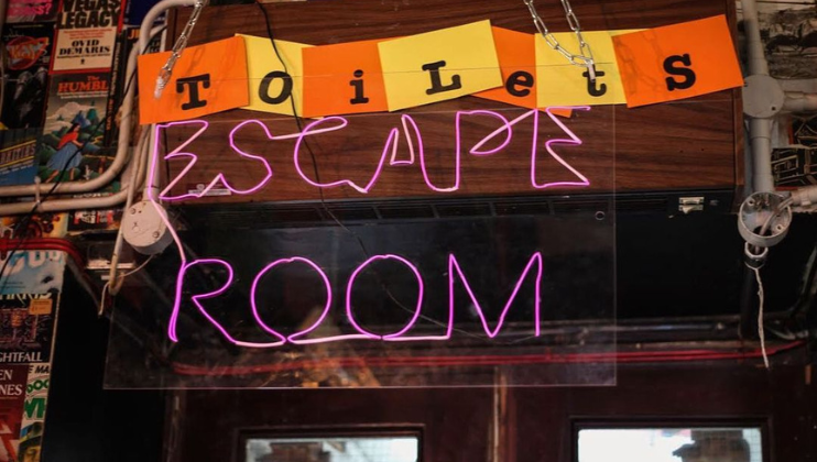 A creatively decorated escape room entrance with a neon pink Escape Room sign. Above it, the word Toilets is humorously split and reassembled on orange and yellow paper square cutouts, adding a playful touch to the ambiance. The backdrop features various posters.
