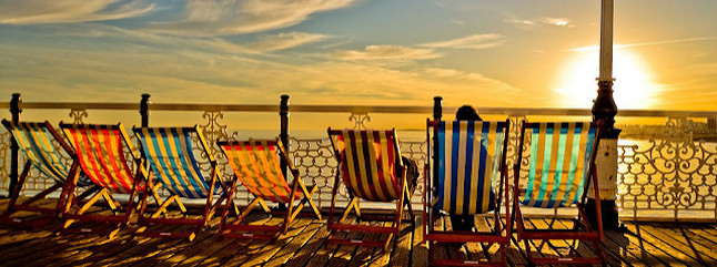 A row of colorful, striped deckchairs on a wooden pier overlook the ocean at sunset. The sky is painted in warm hues, and the sun is partially visible near the horizon, casting a golden glow on the scene.