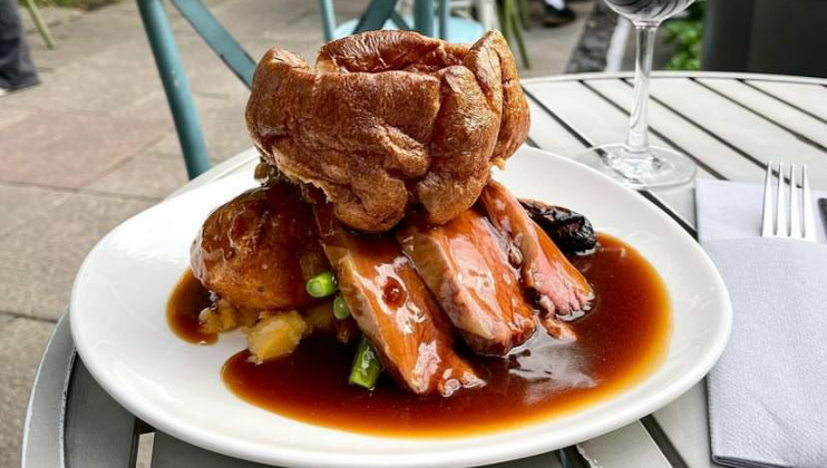 A plate of roast beef slices topped with a large Yorkshire pudding, accompanied by vegetables, roast potatoes, and rich gravy. The dish is placed on an outdoor table next to a glass of red wine and a fork and knife wrapped in a napkin.