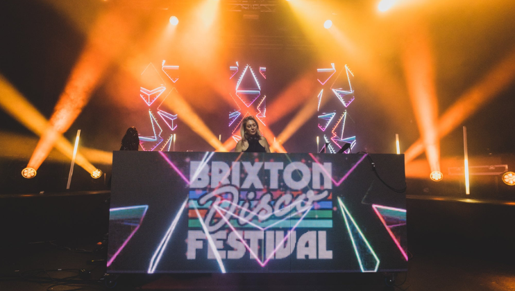 A DJ stands behind a large console with Brixton Disco Festival displayed on it. The background features colorful, illuminated geometric shapes, and beams of light and fog create a vibrant atmosphere. The DJ is surrounded by equipment and spotlights.