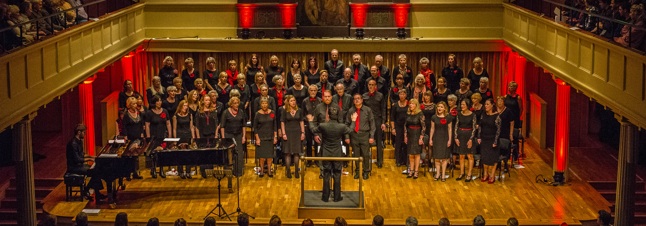 A choir of around fifty people, dressed in black with red accents, stands on a wooden stage performing in a large concert hall with red lighting. The conductor stands in front, facing the choir, while a pianist accompanies them on the left side of the stage.