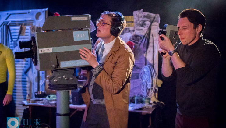 Two individuals are operating old-fashioned broadcasting equipment. One person, wearing a brown coat and headphones, looks focused while adjusting a vintage TV camera labeled RTP. The other person, in darker clothes, operates a smaller, handheld camera.