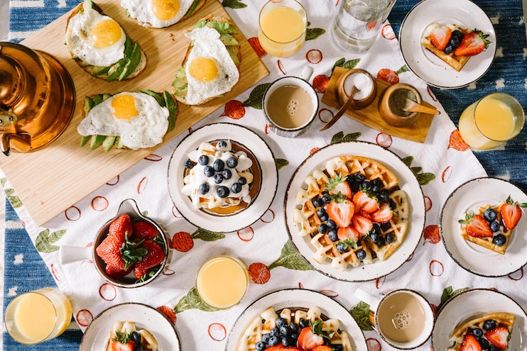 A breakfast spread on a table with a white tablecloth adorned with red and green flowers. The meal includes waffles topped with strawberries and blueberries, sunny-side-up eggs, avocado toast, bowls of strawberries, glasses of orange juice, and a kettle.