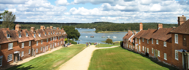 A wide path lined with neatly trimmed grass runs through an old village with symmetrical rows of brick houses on either side. At the end of the path, a lake with boats and a forested area are visible in the background under a partly cloudy sky.