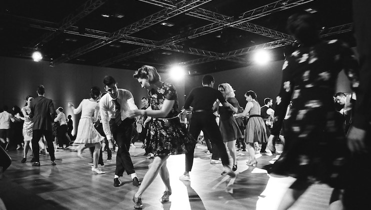 A lively scene of people swing dancing in a spacious hall with an industrial-style ceiling. The dancers, wearing mid-20th-century styled clothing, are engaged in various dance moves. The black-and-white image captures the energy and joy of the event.