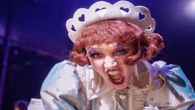 A performer dressed in a whimsical costume with heart-shaped cutouts and exaggerated makeup, including dramatic eye shadow and red lipstick, expresses a fierce or intense emotion on stage against a dark, blurred background.