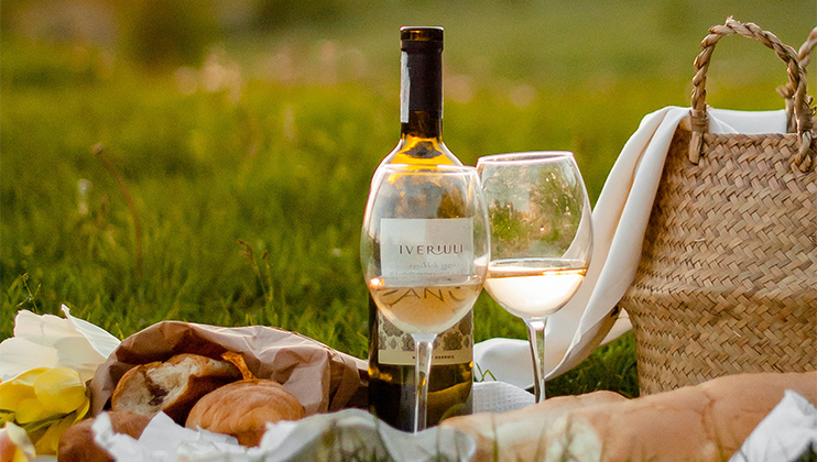 A scene of a picnic on grass features a bottle of white wine labeled “Iveruli” and a filled wine glass. Beside them are a woven basket, a wrapping paper with bread, a fabric cloth, and blurred flowers in the background.
