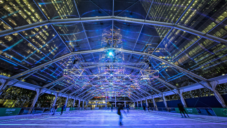 Indoor skating rink with a clear roof and colorful illuminated lights creating patterns above. Blurred figures of skaters glide on the ice, and surrounding buildings are visible through the transparent structure, adding a vibrant urban backdrop.