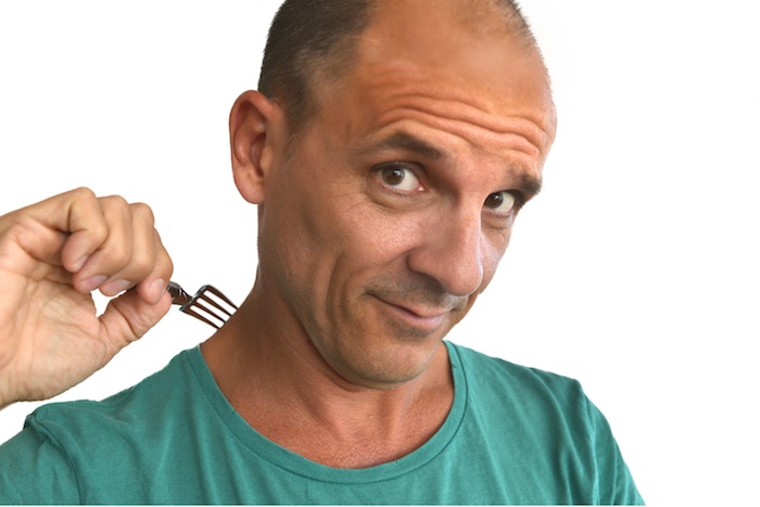 A bald man with a green t-shirt is playfully holding a small fork close to his neck, with a mischievous expression on his face. The background is plain white.