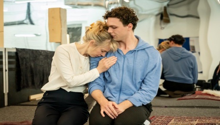 Two people sit closely on a carpet. A woman, wearing a white blouse, leans her head on the shoulder of a man in a light blue hoodie with his eyes closed. In the background, there are mirrors and blurred figures, suggesting a rehearsal or contemplative setting.
