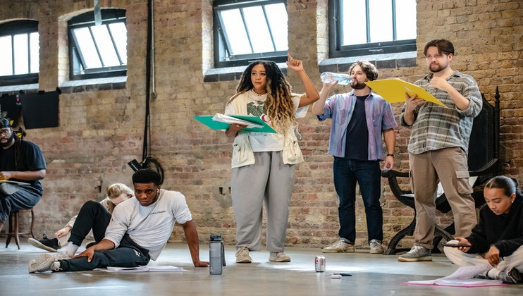 A group of actors in casual clothes are rehearsing in a spacious room with brick walls and tall windows. One actor, holding a green binder, is speaking with her hand raised. Others are either sitting on the floor or standing, some holding scripts and water bottles.