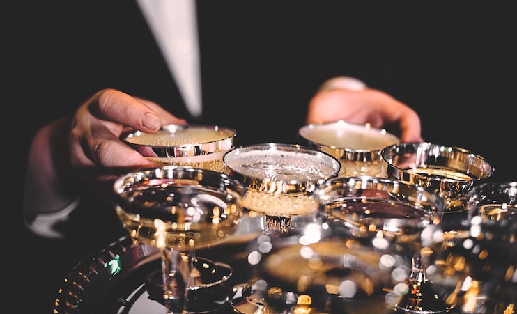 A person in formal attire holding a tray filled with multiple silver champagne saucers, with champagne bubbling inside. The background is dark, making the tray and glasses stand out prominently.