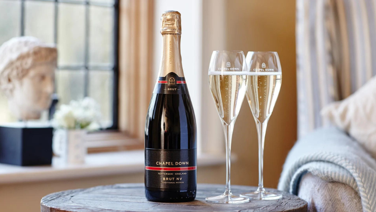 A bottle of Chapel Down Brut NV sparkling wine is placed on a round wooden table, accompanied by two filled flutes. The background features a window with a classical sculpture and a potted plant, adding elegance to the scene.