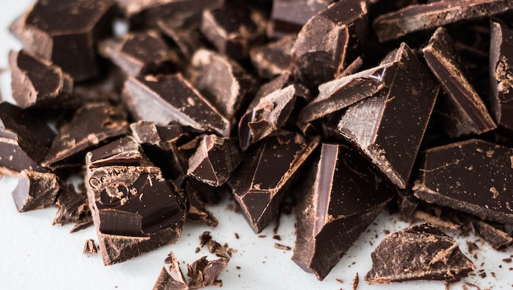 Close-up of scattered chunks of dark chocolate on a light surface. The chocolate pieces are irregularly shaped and vary in size, showcasing a rich, glossy texture with some crumbs around them.