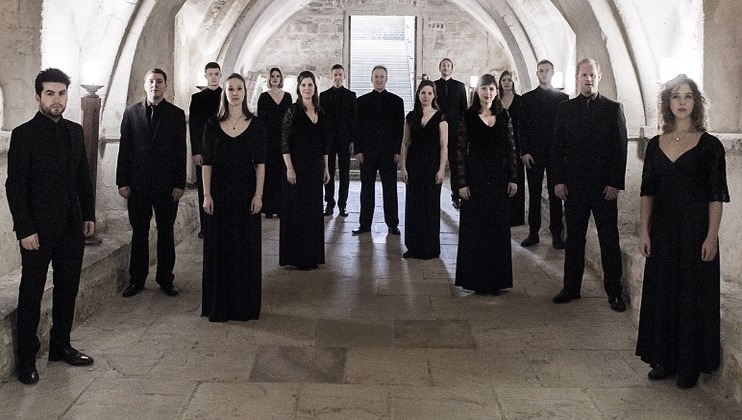 A group of 15 people, dressed in black formal attire, stand evenly spaced in two rows along a stone corridor with arched ceilings. They are positioned facing the camera, under a soft, natural light that filters through a window in the background.