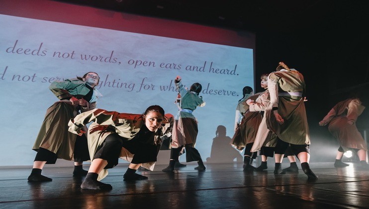 A group of performers on stage, dressed in uniform-like costumes, engaged in an intense and expressive dance. The background features a large projection with text that reads, deeds not words, open ears and heard, and additional partial phrases.