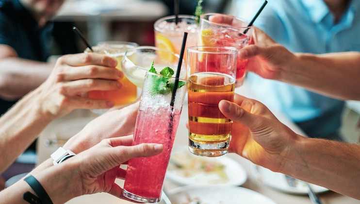 A group of people raise their glasses in a toast. The drinks include colorful cocktails garnished with mint and lime, as well as a glass of beer. The background is blurred, focusing on the hands and glasses in celebration.