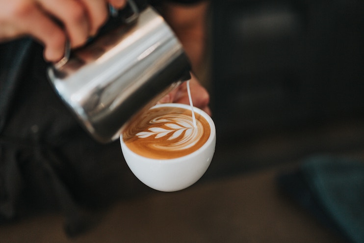 A barista is pouring steamed milk from a metal pitcher into a cup of coffee, creating a leaf-shaped latte art design on the surface. The cup is held in one hand, while the other hand skillfully pours the milk.