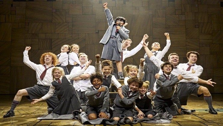 A group of children performing on stage, dressed in school uniforms with grey sweaters and shorts or skirts. One child stands triumphantly on another's shoulders, raising a fist, while the others cheer enthusiastically around them. The background is a wooden mosaic.