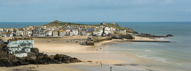 A panoramic view of a coastal town with sandy beaches meeting the blue sea. Numerous houses and buildings are clustered on the shoreline with a breakwater extending into the water. Small boats are scattered along the beach, and the sky is partly cloudy.