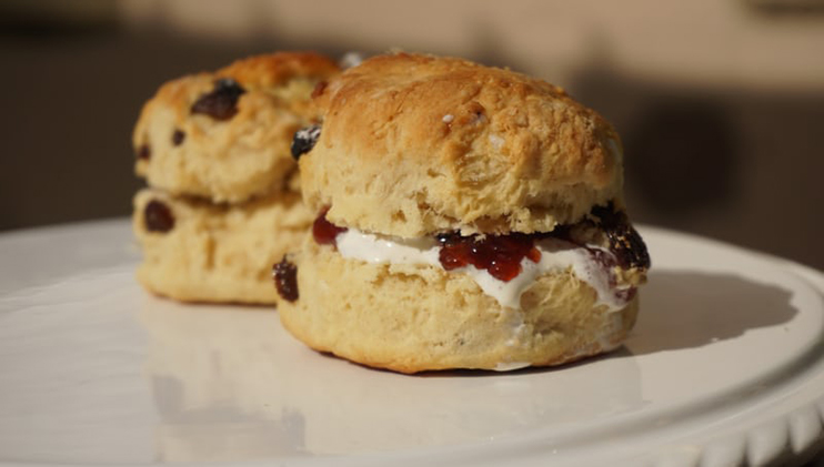 Two freshly baked scones with a golden-brown crust sit on a white plate. Each scone is split and filled with a layer of clotted cream and strawberry jam, with bits of dried fruit visible in the dough. The background shows a softly blurred, neutral-colored setting.