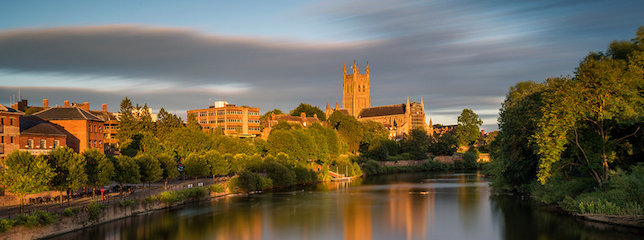 A picturesque view of a town with a large cathedral surrounded by trees and buildings. Below, a tranquil river reflects the greenery and structures along its banks, while soft, elongated clouds streak across the blue sky.
