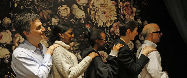 Five people standing in a line, each with their hands on the shoulders of the person in front of them. They appear to be participating in an activity or game. The background features a dark, floral-patterned wall.