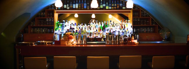 A dimly lit, cozy bar with a variety of colorful bottles on shelves behind the counter. The bar features several glowing pendant lights. Four stools with light cushions line the counter, and there are various cocktail shakers and garnishes on the bar top.