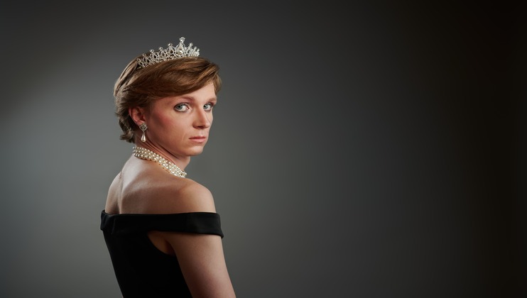 A person with short hair wears a diamond tiara, pearl earrings, and a pearl necklace. They are dressed in an off-the-shoulder black gown, looking over their shoulder against a gradient dark background.