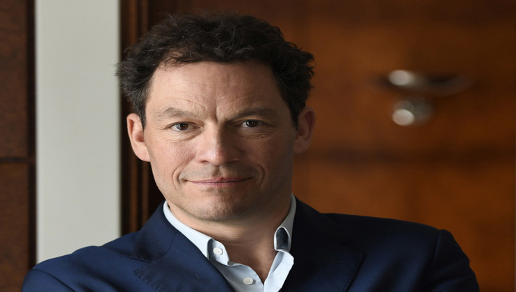 A man with short curly dark hair, wearing a blue blazer over a light blue shirt, smiles slightly. He is posed in front of a blurred background that appears to have wood-paneled walls. His arms are crossed, and he is looking directly into the camera.