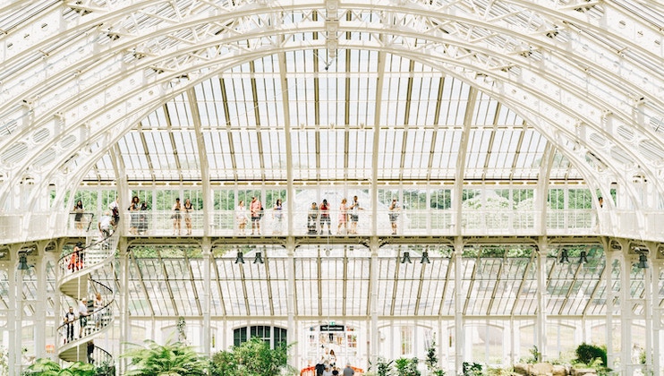 A bright, spacious greenhouse with a large arched glass roof and intricate white metal framework. Visitors are walking along an upper balcony while lush green plants flourish below. A spiral staircase on the left connects the levels.