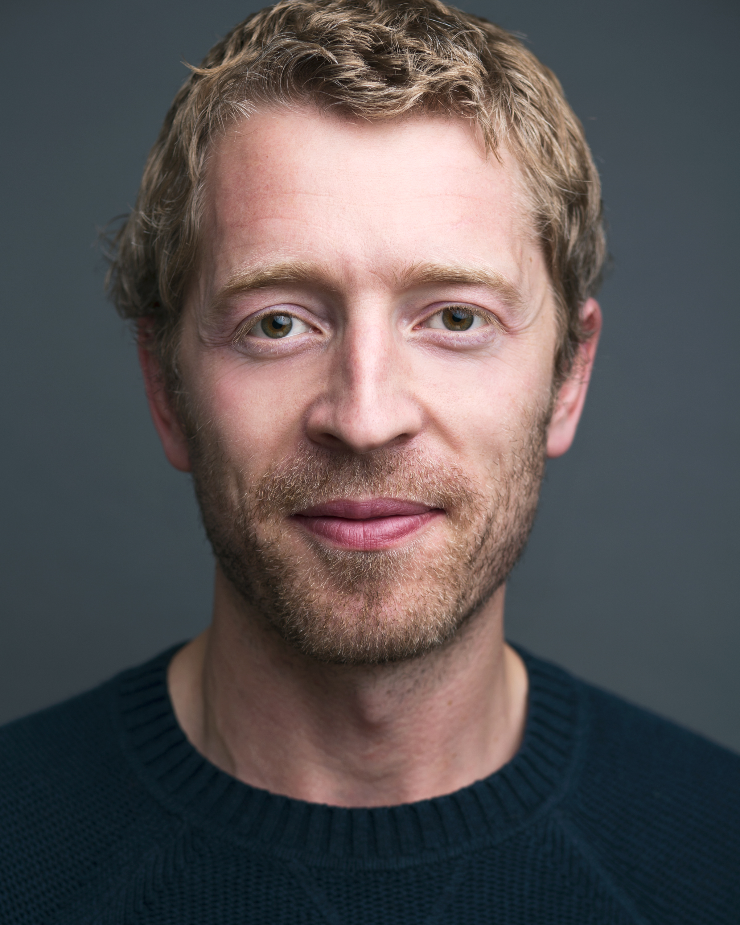 A man with short, light brown hair and a beard is looking directly at the camera. He is wearing a dark blue sweater and has a neutral expression. The background is plain and gray.